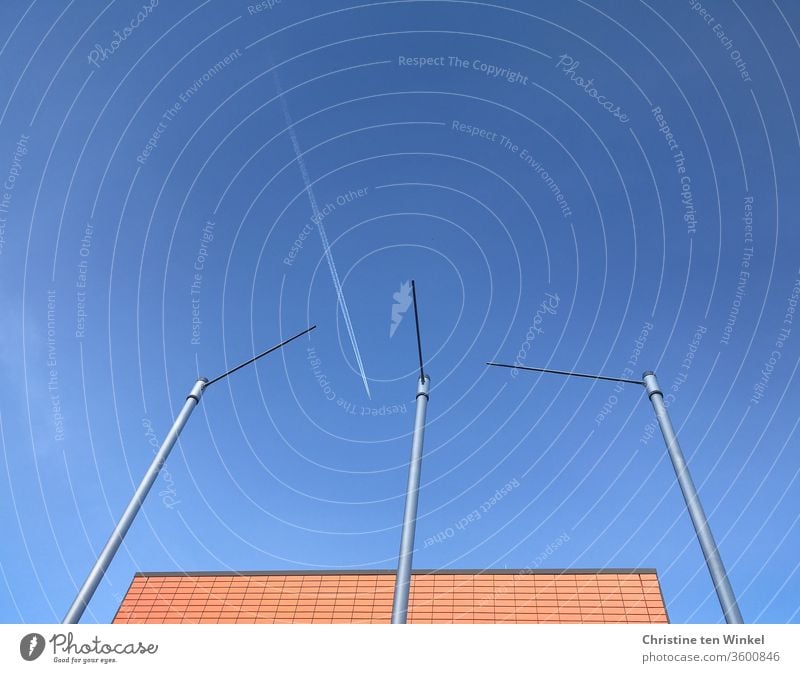 white contrail between empty flagpoles, blue sky and partial view of an orange building Vapor trail Airplane Flying Sky Blue sky Summer Cloudless sky Flagpoles