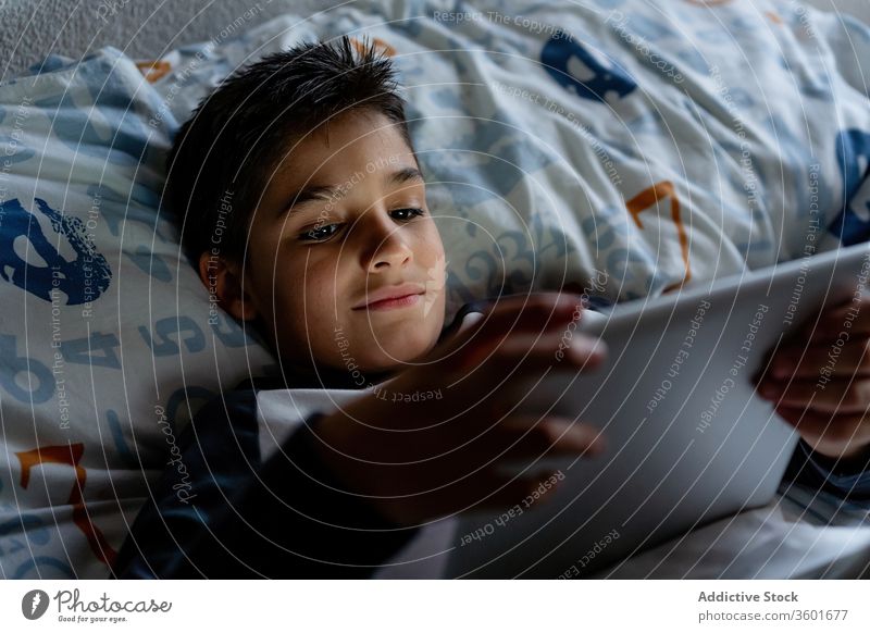 Delighted boy using tablet in bedroom during weekend kid blanket night watching cheerful pajama home adorable device gadget child internet browsing cozy rest