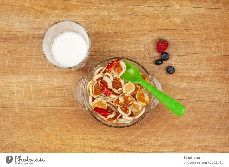 Mini pancakes with strawberries and blueberries in a glass bowl are on a wooden table, next to a glass of milk. The view from the top, place for text breakfast