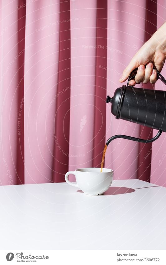 Woman's hand holding black metal pot and pouring coffee or tea into white cup in front of pink drapery, selective focus beverage morning drink mug breakfast