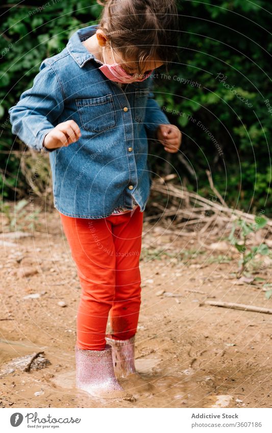 Curious girl surgical mask playing in mud puddle child having fun curious kid rubber boot cute dirty water wet adorable childhood joy activity playful ripple