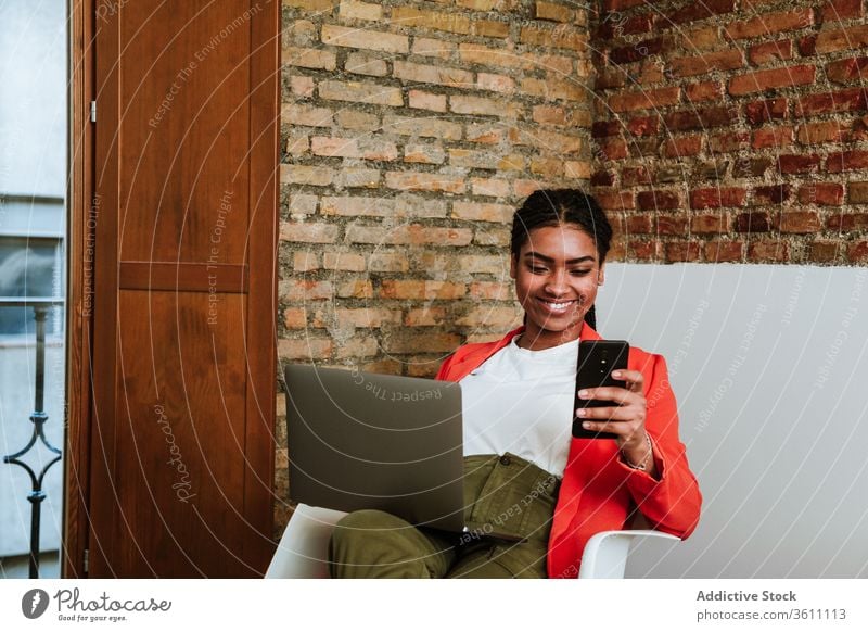 Smiling modern black woman with gadgets in armchair business laptop cheerful smartphone online elegant computer loft design interior freelance happy device