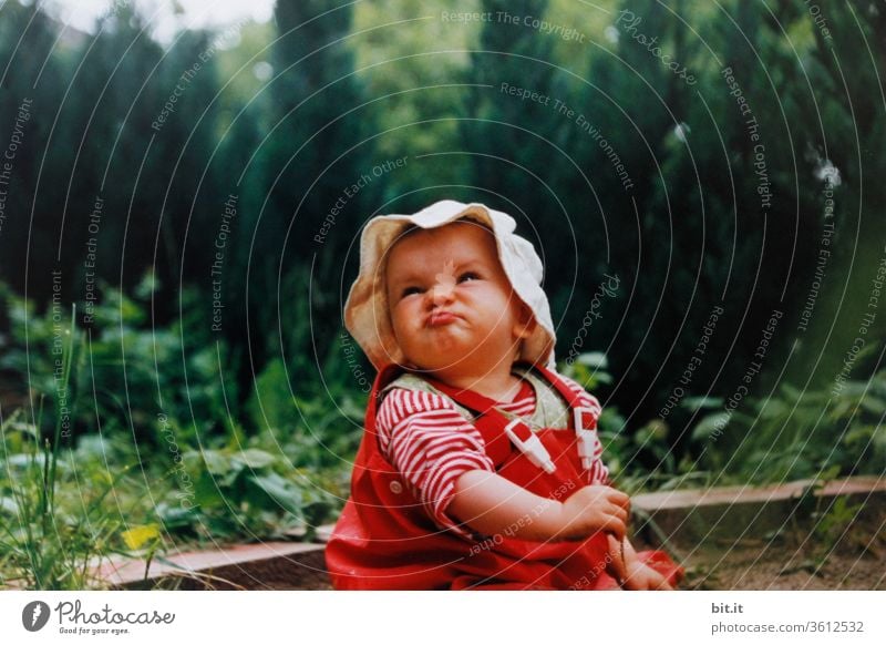 Little girl is sitting with red mud pants and a cap with frills, in the sandbox from the garden, with a green hedge in the back. The child pulls a sweet, funny, cute face and looks funny, curious, interested, up into the air. Rainy day at home.