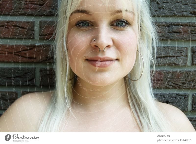 Portrait of a young blonde woman in front of a brick wall girl Young woman already smile Blonde long hairs Earring blue eyes Skin youthful 19 18-20 years