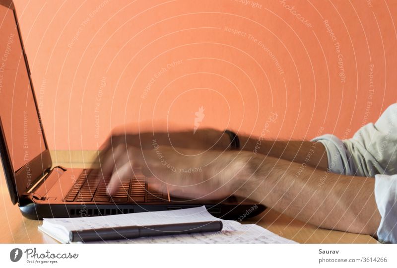 A motion blurred close up shot of the fingers of a young adult Working from Home on a laptop kept on a wooden desk next to a pen and a notepad. work from home