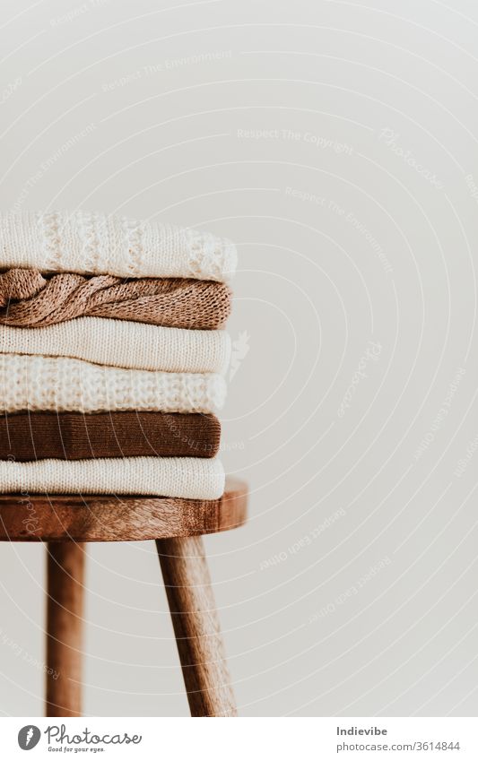 Women fashion composition. Warm winter female woollen knitted sweaters, pullovers stack on a wooden stool at white background. Modern clothes concept for magazine, blog, social media.