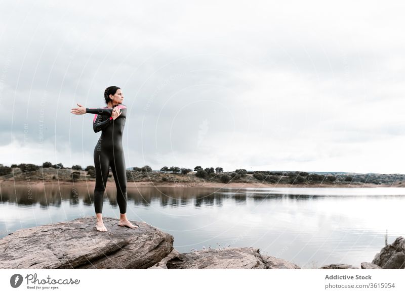 Calm woman stretching on rock on lake shore nature warm up diver wetsuit harmony calm overcast water sports wellness flexible barefoot slim healthy energy