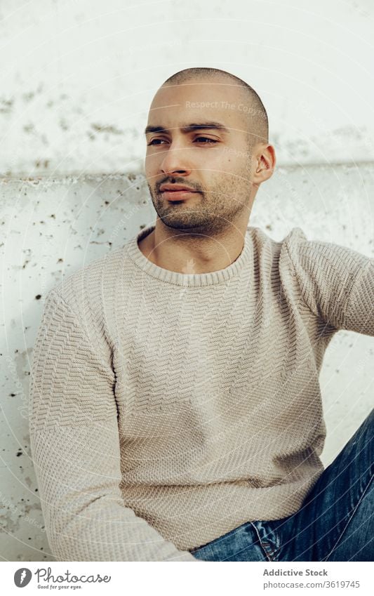 Stylish young man sitting on concrete wall style trendy pensive hipster modern street style thoughtful portrait contemplate unshaven guy jeans casual ethnic