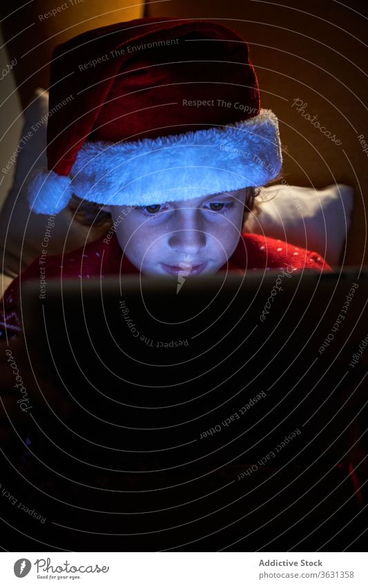 Happy kid enjoying new tablet on Christmas eve at home boy christmas santa hat holiday toothy smile internet online using gadget device reflection child