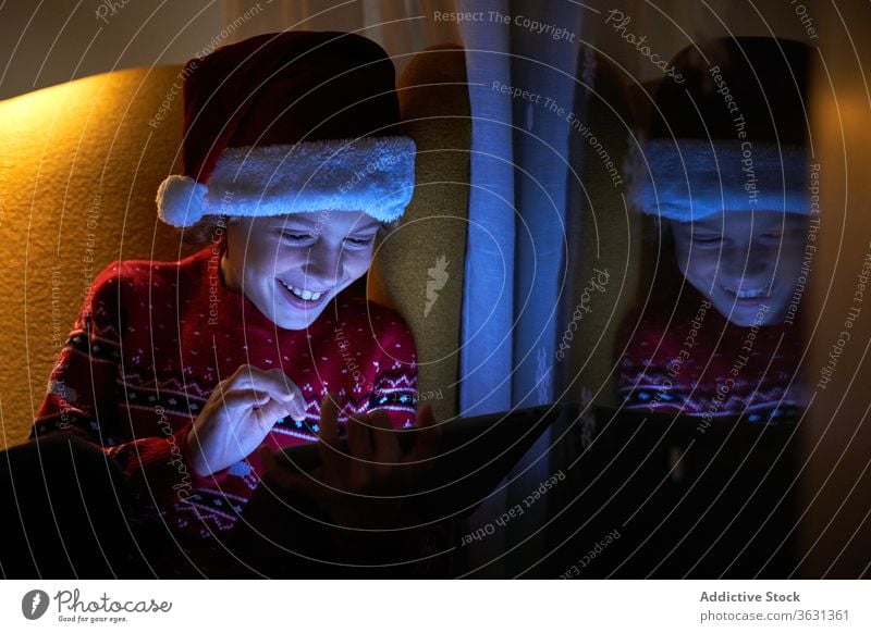 Happy kid enjoying new tablet on Christmas eve at home boy christmas santa hat holiday toothy smile internet online using gadget device reflection child