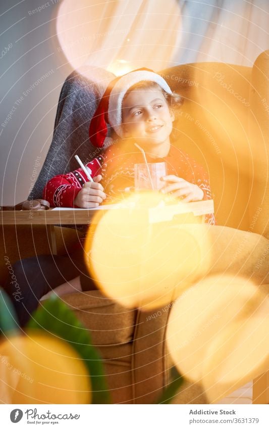 Happy boy in Christmas wear sitting in cozy armchair santa hat christmas holiday pencil happy comfort domestic sweater glad cheerful comfortable bokeh positive