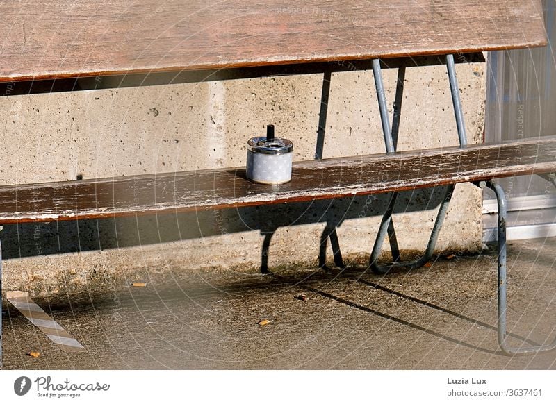 A smoking area in the sun: brown wooden bench, stars on the ashtray, lots of sun and long shadows on the floor Ashtray asterisk Wooden bench seat Old Brown