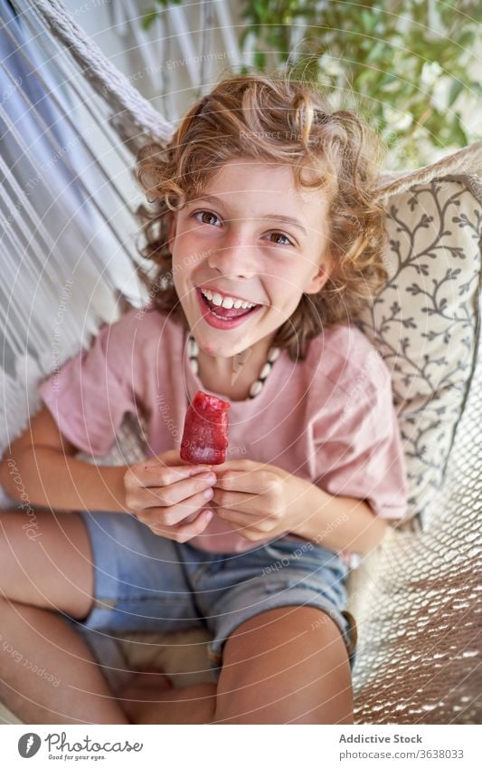 Content boy enjoying delicious fruit ice lolly sitting in hammock eat kid weekend legs crossed barefoot mouth opened idyllic childhood comfort rest harmony