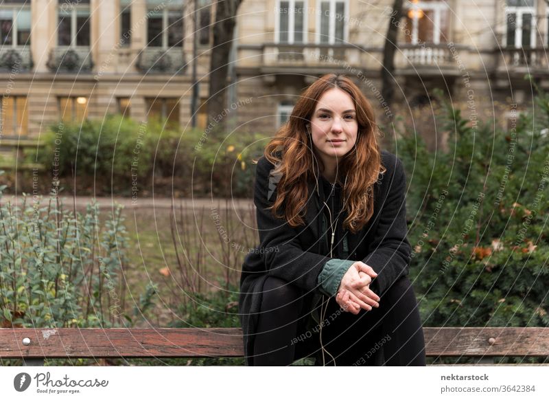 Young Woman Looking at Camera Seated on Bench caucasian ethnicity woman female earphones audio music listening brown hair real life model real person urban