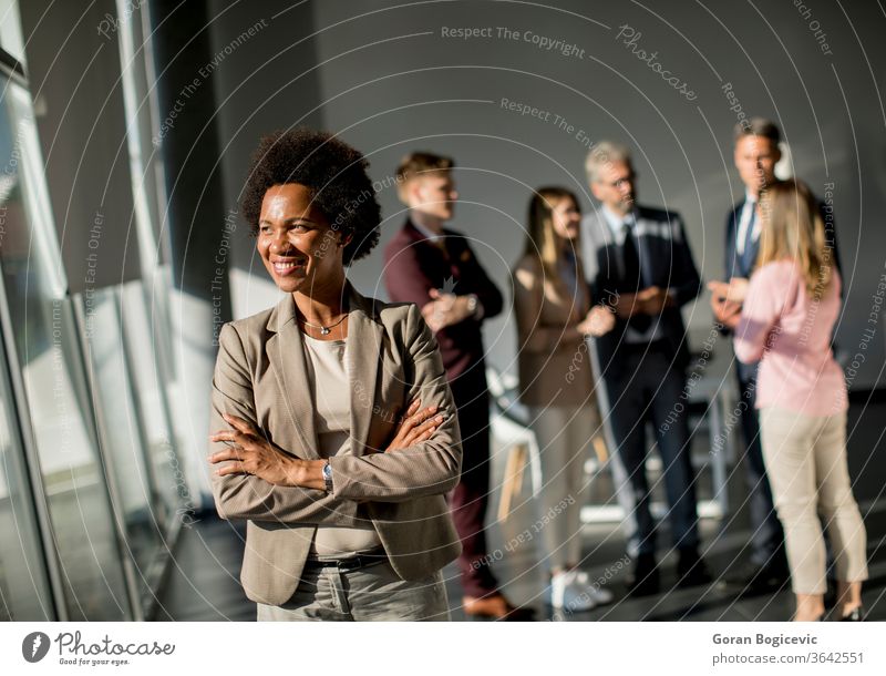 Professional black woman standing in office with folded arms and confident expression as other workers hold a meeting in background business teamwork career