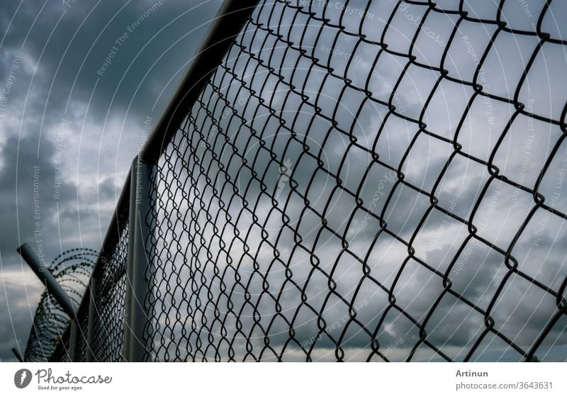 Military zone mesh fence. Prison security fence. Looking up view of barbed wire security fence with storm sky and dark clouds. Razor wire jail fence. Barrier border. Boundary security wall.