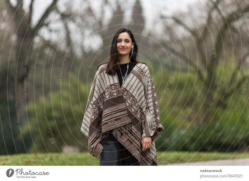 Young woman with tanned complexion wearing poncho in park and smiling. young woman Indian ethnicity middle eastern ethnicity earphones medium shot audiobook