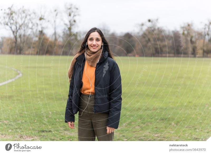 Young Indian woman smiling on public park lawn. Front view. young woman Indian ethnicity music earphones medium shot audiobook listening focus on foreground