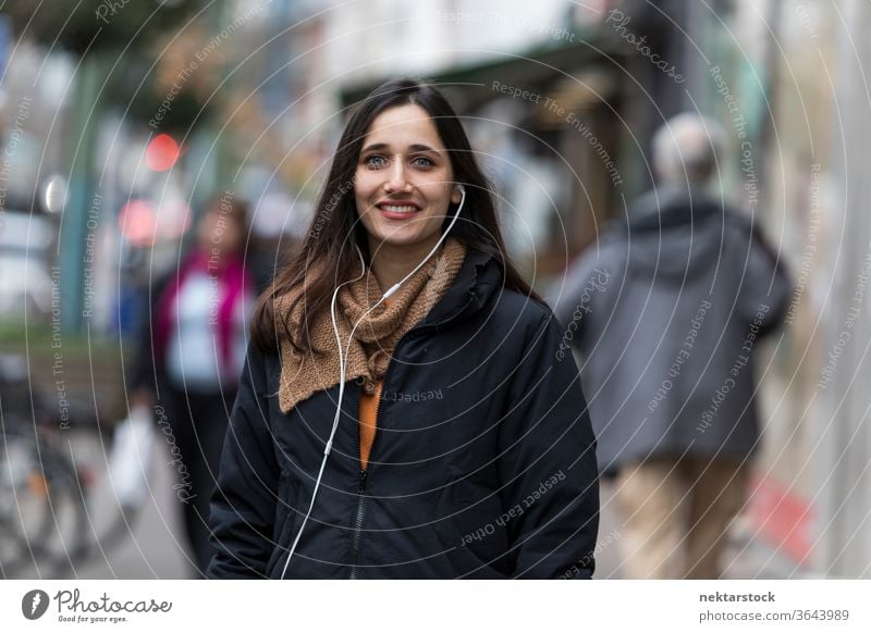 Young Indian woman listening to earphones and smiling toothy smile on street. young woman Indian ethnicity music medium shot audiobook focus on foreground