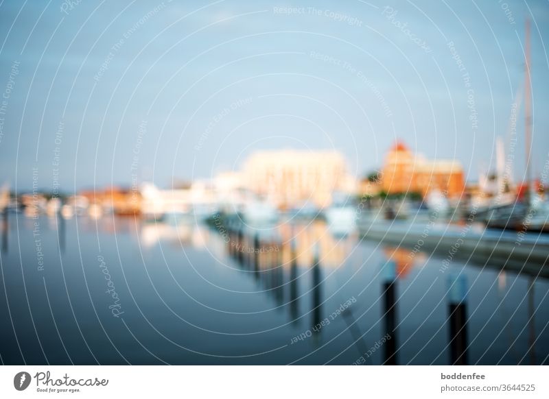 dreamy, blurred view of the skyline of Barth harbour, evening atmosphere with blue sky and buildings illuminated by the evening sun, reflected in the water. In the foreground diagonally to the back middle there are poles for leashing boats