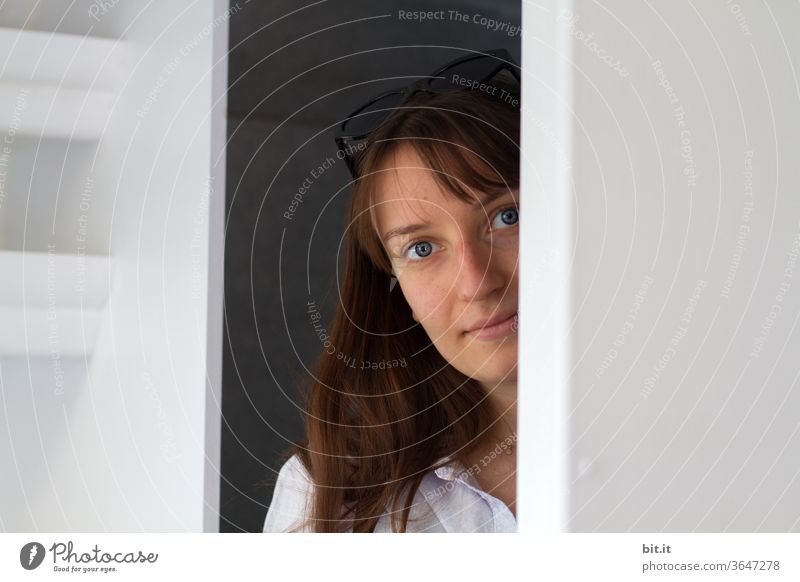 Young woman with sunglasses on her head, looking out behind a white wall. Pretty, roll-eyed woman looks curiously around the corner and smiles gently into the camera. Beauty photography between two walls, at home in a bright room of the apartment.