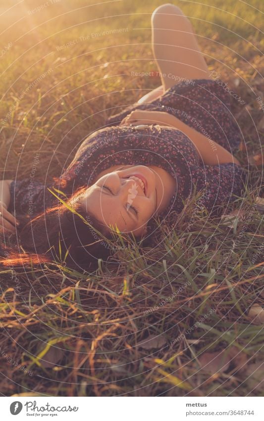 Happy girl is lying down in autumnal grass and laughing backlit by sunset sky activity back view bare legs communication crossed legs crosslegged female green