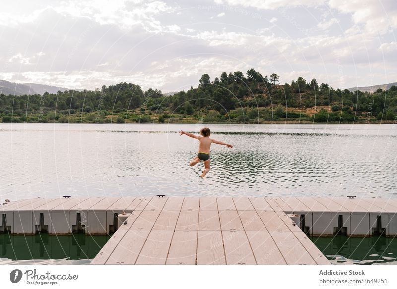 Delighted boy jumping in lake from pier having fun water moment kid pond childhood swimwear quay wooden weekend summer vacation relax joy rest together activity