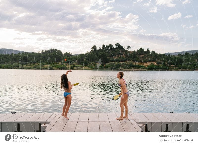 Happy sisters playing beach tennis on pier lake girl active enjoy holiday having fun bikini summer ball racket teenage quay wooden vacation happy pond delight