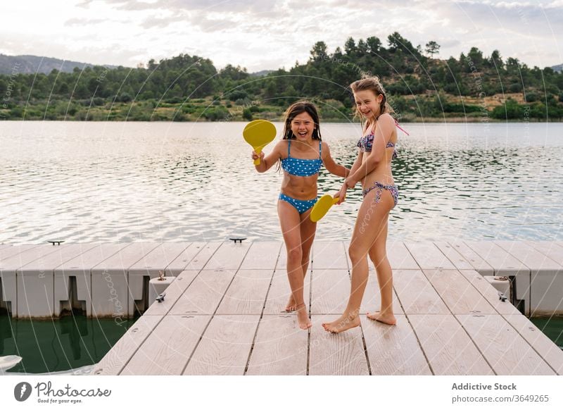 Happy girls playing beach tennis on pier lake sister active enjoy holiday having fun bikini summer ball racket teenage quay wooden vacation happy pond delight