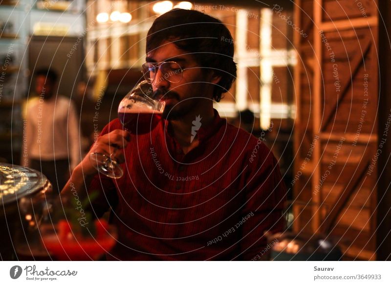 Young Man wearing red shirt while drinking Red Wine from a goblet at a bar indoors with colorful lights all around him. lifestyle celebration alcohol wine