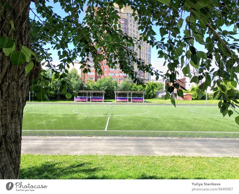 Empty green soccer field near a school and the apartment house in Russia. Stadium and football field with empty spectator seats. Keeping fit and exercising outdoor. Urban view in a frame of leaves.