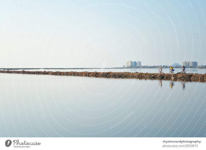 Reflections of 3 pedal bike riders shimmer in a still lagoon mediterranean landscape people cyclists water reflection mirror image sparse empty tranquil calm