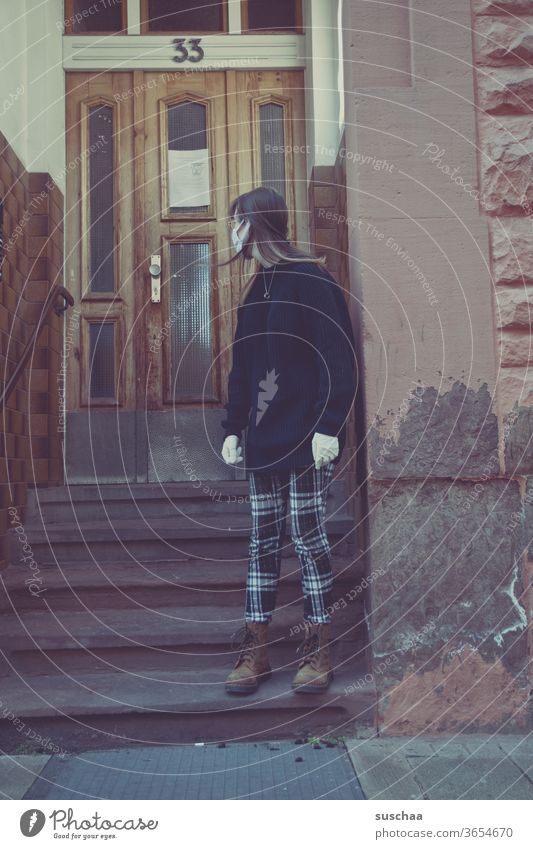 teenager with face mask and latex gloves is standing in front of an apartment building and looks over the shoulder direction door with the number 33