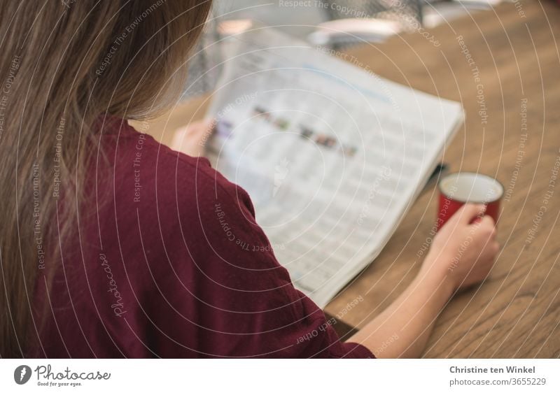 Print I A young woman is sitting outside at a table reading the newspaper. With her right hand she is holding a red coffee mug. A smartphone is lying on the table, almost covered by the newspaper.