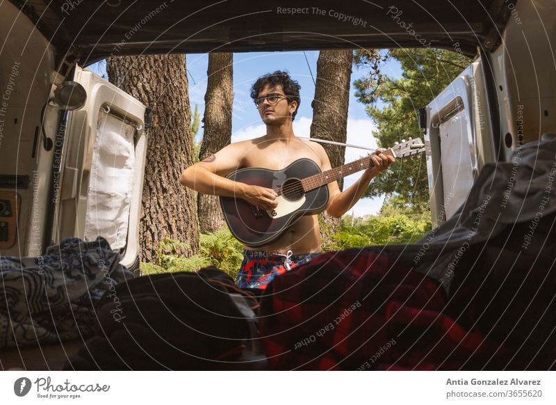 Guy playing adventure guitar in the forest (camping) seen from the inside of a minivan Relaxation Guitar recreation tourism journey trip outside young smile