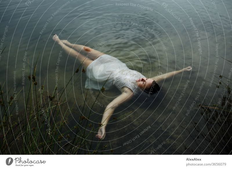 Young woman in white summer dress lies in the water near the shore of a forest lake Woman girl plants green Nature Dress Barefoot barefoot Slim Athletic Legs