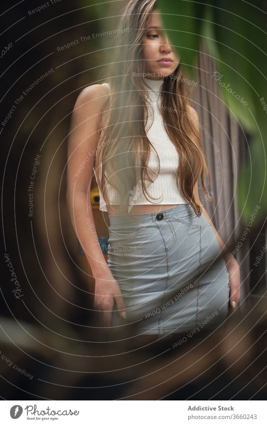 Graceful young woman in trendy outfit standing near curtains thoughtful pensive room green plant leaf upset alone calm dreamy female slim long hair mini skirt