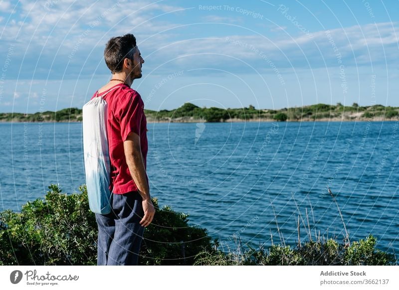 Thoughtful young man enjoying seascape against blue sky relax hill admire calm peaceful recreation nature harmony tranquil male casual yoga mat bag serene rest