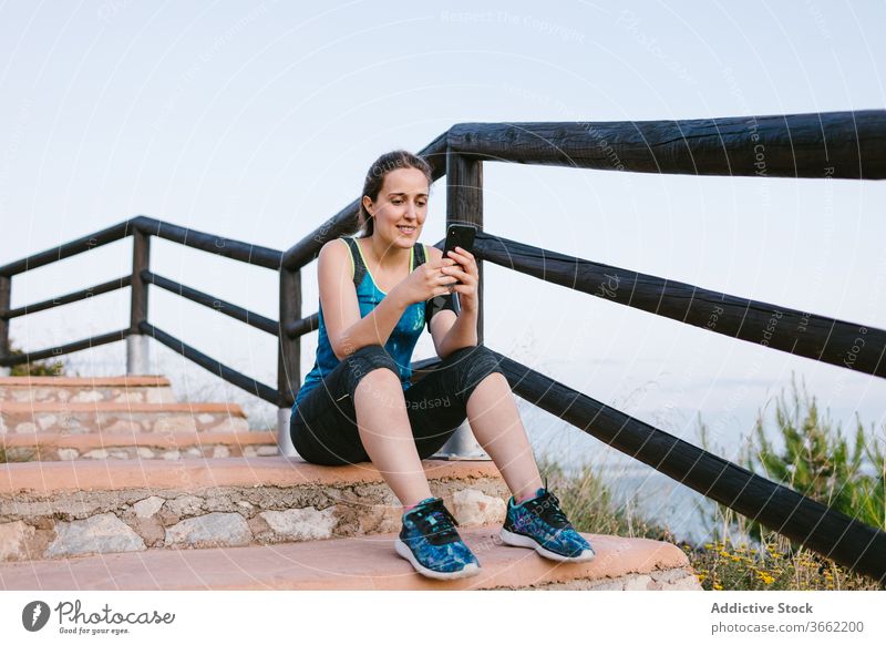 Sportswoman sitting on stairs with mobile phone near sea sportswoman break rest alone sportswear athlete staircase healthy fit wellbeing ocean summer activewear