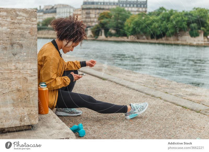 Sportswoman in headset using social media on smartphone after workout sportswoman headphones chatting internet break embankment river gadget device online