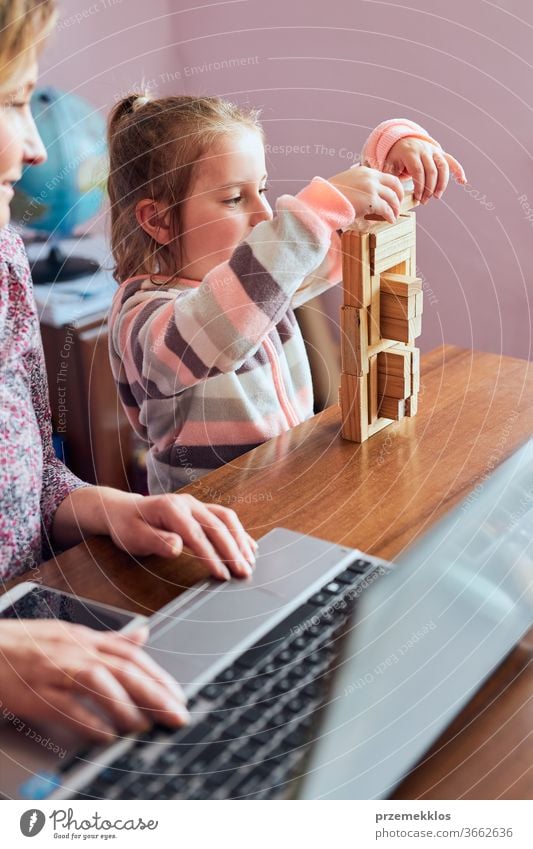 Woman mother working doing her job remotely during video chat call stream online course webinar on laptop from home while her daughter playing with bricks toy. Woman sitting at desk in front of computer looking at screen