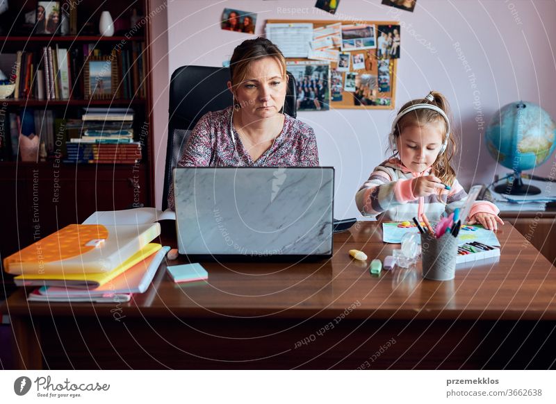 Woman mother working doing her job remotely during video chat call stream online course webinar on laptop from home while her daughter making drawings playing around and listening to stories. Woman sitting at desk in front of computer looking at screen