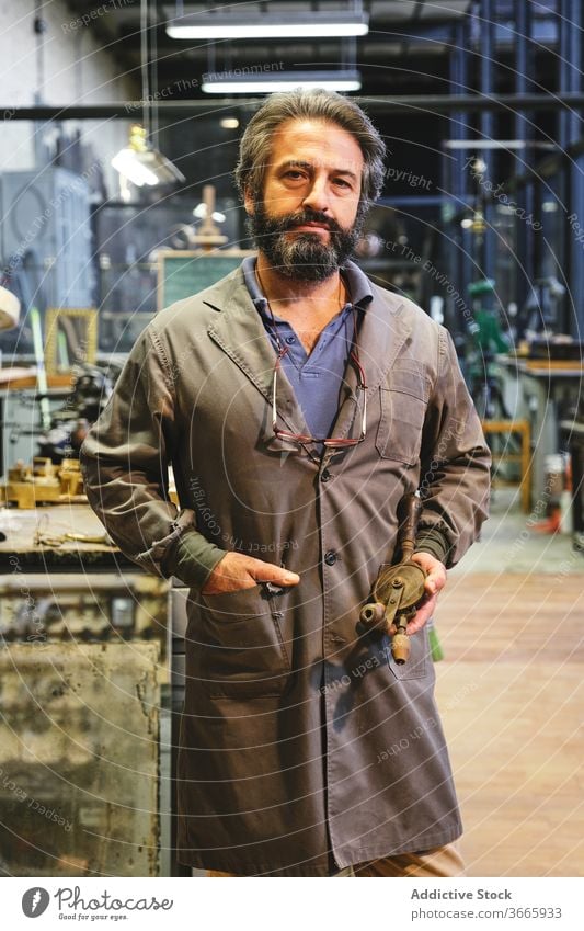 Serious ethnic craftsman standing with golden object in workshop jeweler handicraft serious hand in pocket uniform small business studio art goldsmith workspace