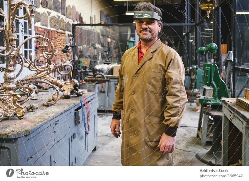 Smiling artisan in headband magnifier standing near golden item jeweler uniform artwork craft profession small business workshop smile studio man goldsmith robe