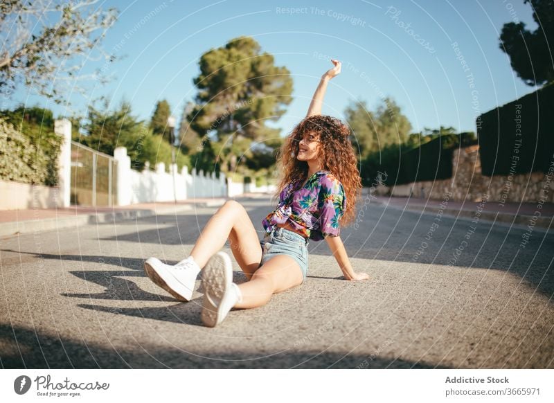 Gorgeous woman sitting with raised arm on asphalt road arm raised toothy smile eyes closed happy lifestyle excited harmony blue sky carefree tree cheerful