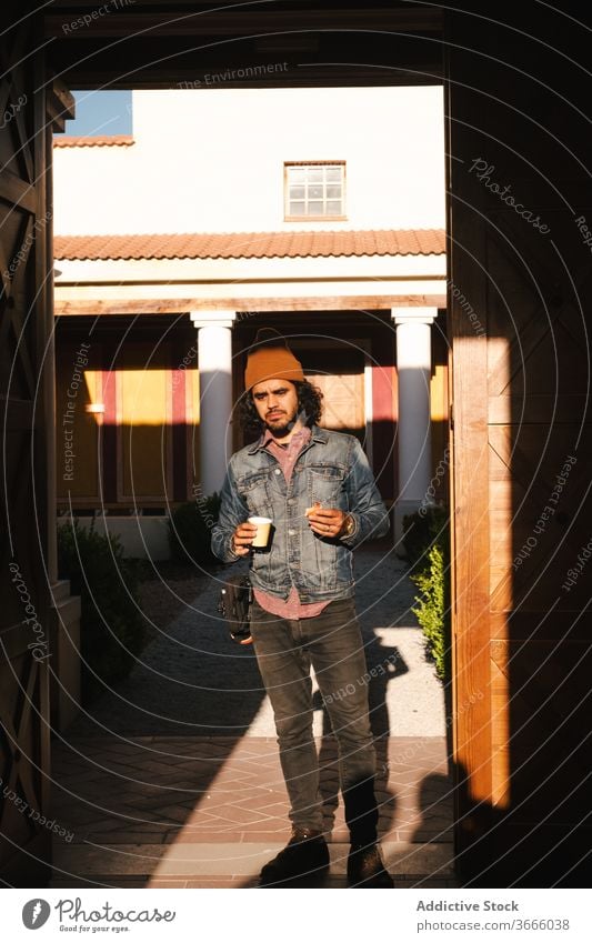 Man with paper cup against building with pillars man refreshment drink positive street exterior hipster coffee break content outfit colorful evening sunlight