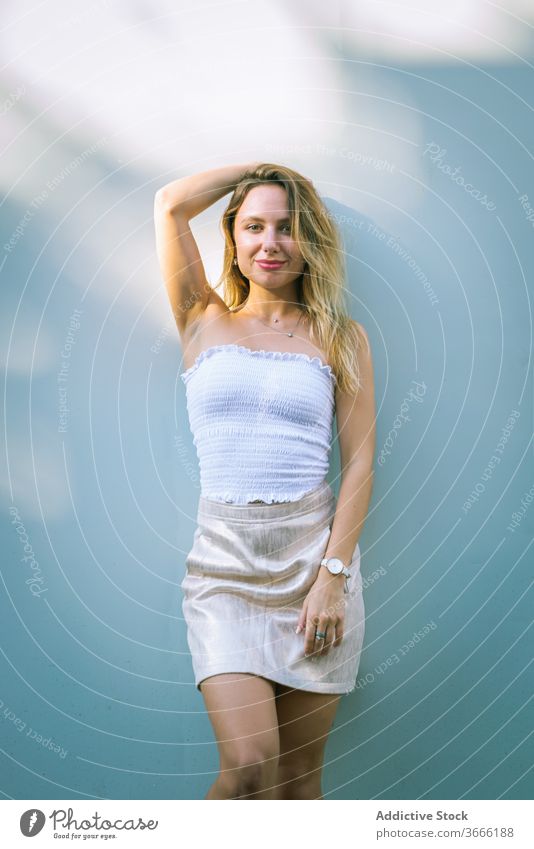 Positive young blonde standing near white wall on sunny day woman confident smile sunlight shadow style portrait touch head daytime positive female slim summer