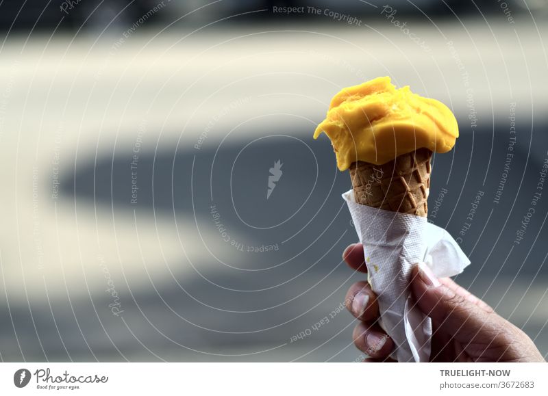 If the weather is nice and hot - all children want ice cream: Mango - Mango! Schleck - Schleck! A woman's hand holding an ice cream cone with a ball of yellow mango ice cream with a paper napkin in front of light and shadow on the street