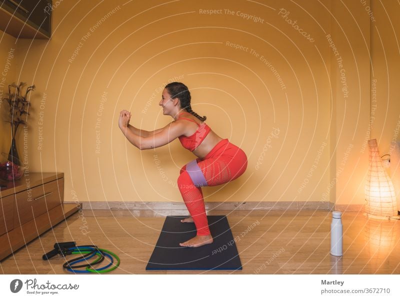 Brunette girl enjoying exercising squatting working buttocks in the living room with some tapes. Beautiful caucasian girl exercising at home by the covid. woman