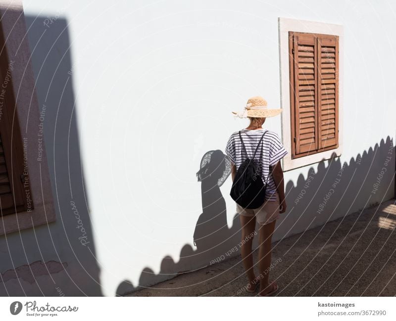 Rear view of beautiful blonde young female traveler wearing straw sun hat sightseeing and enjoying summer vacation in an old traditional costal town at Adriatic cost, Croatia.